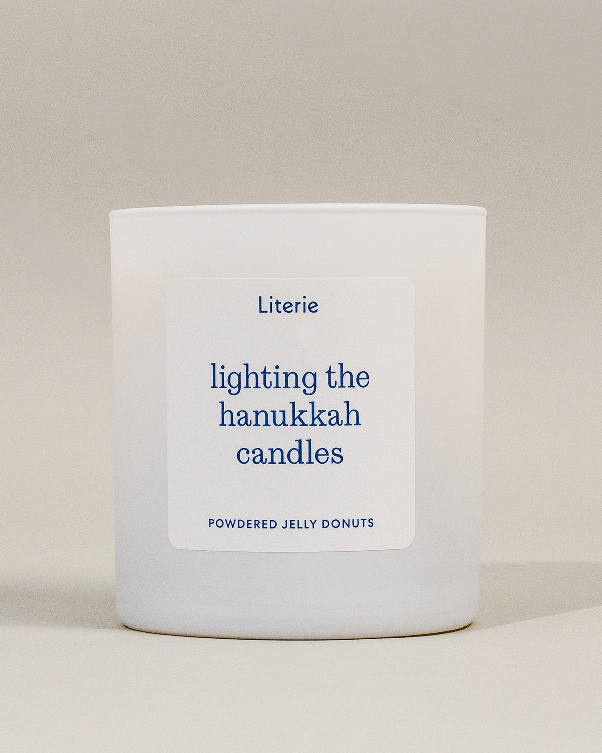 A white glass candle jar with a white rectangular label featuring blue text that reads 'Literie lighting the hanukkah candles' and 'POWDERED JELLY DONUTS' at the bottom, set against a neutral beige background.