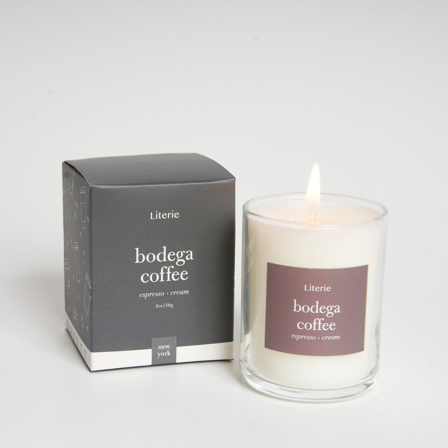 A candle with a lit flame, next to its gray packaging box, labeled 'bodega coffee'.