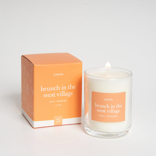 A candle with a white wax and a label that reads 'brunch in the west village', accompanied by a box with matching design and text.