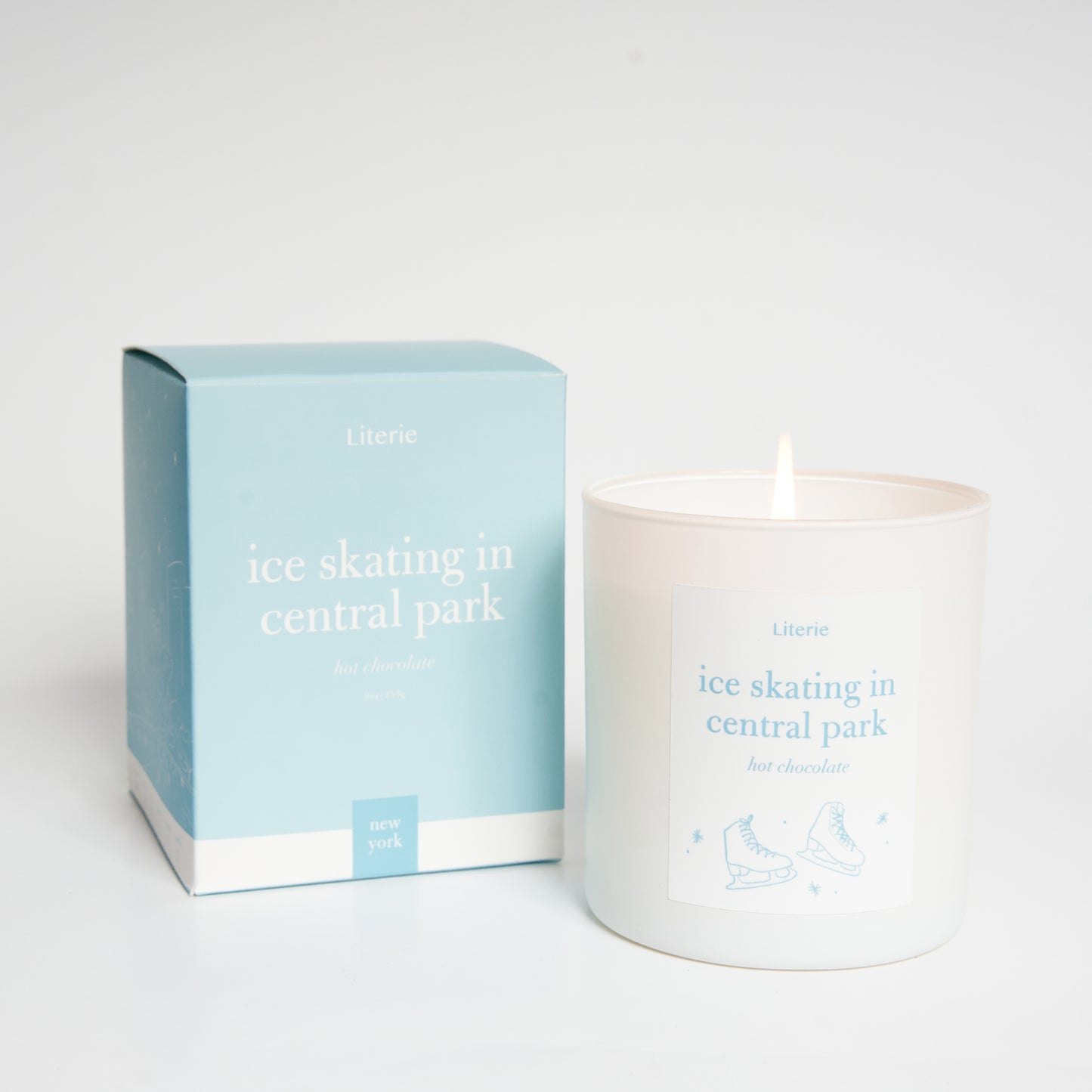 A candle with a label that reads 'ice skating in central park' alongside its packaging box, both featuring a light blue color scheme with text and decorative elements.