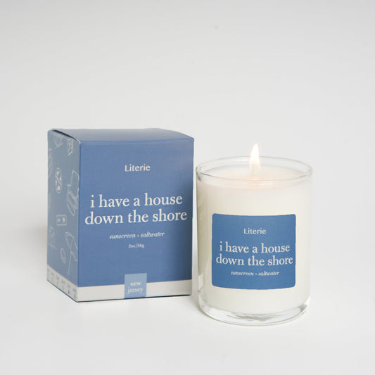 A Literie branded candle with the title 'i have a house down the shore' displayed on its clear glass container, alongside its blue packaging box with the same title.