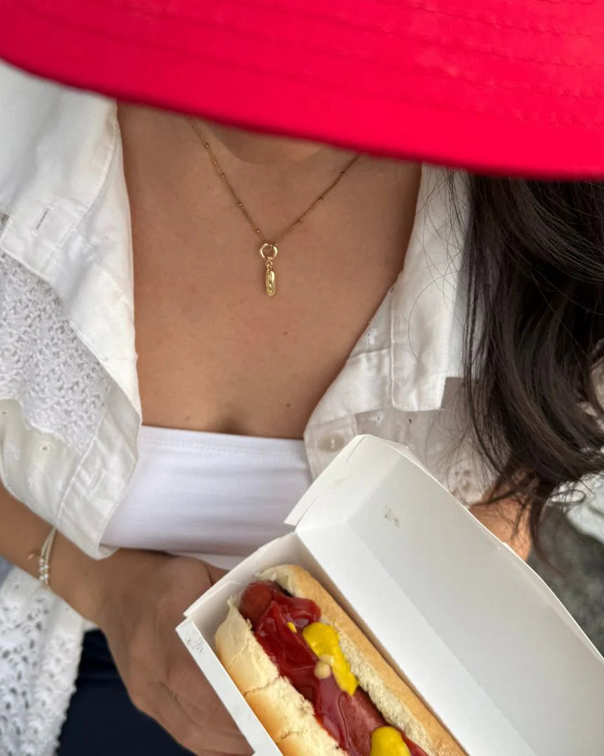 Hotdog charm on necklace with hotdog being held by a woman with a red hat.