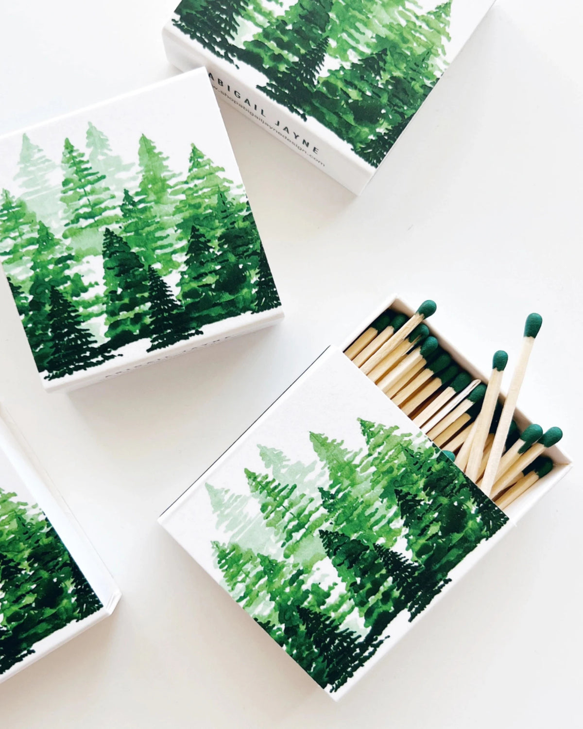 A set of white matchboxes decorated with a green watercolor pine forest pattern, with one open box showing wooden matches with dark green tips on a clean white background.