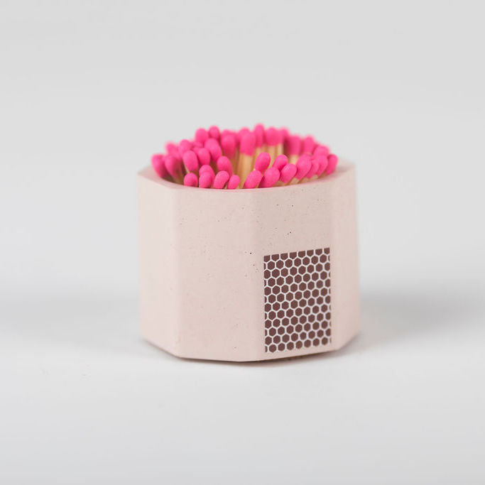 A pink concrete match holder with a hexagonal design and cork bottom, filled with pink matches, on a white background.
