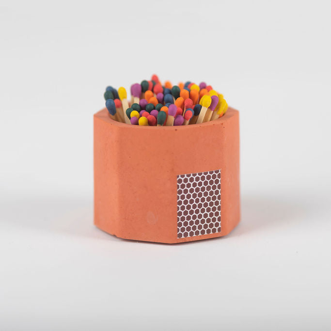 A orange concrete match holder with a hexagonal design and cork bottom, filled with rainbow matches, on a white background.
