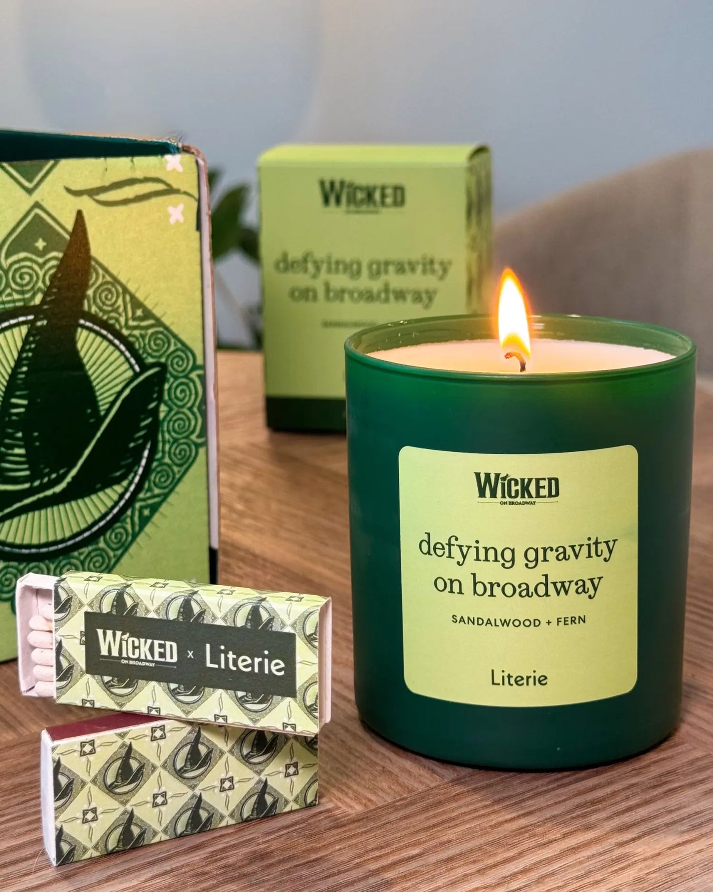 A lit WICKED 'defying gravity on broadway' candle in a dark green glass jar sits on a wooden table next to its patterned packaging box and two wrapped matches.