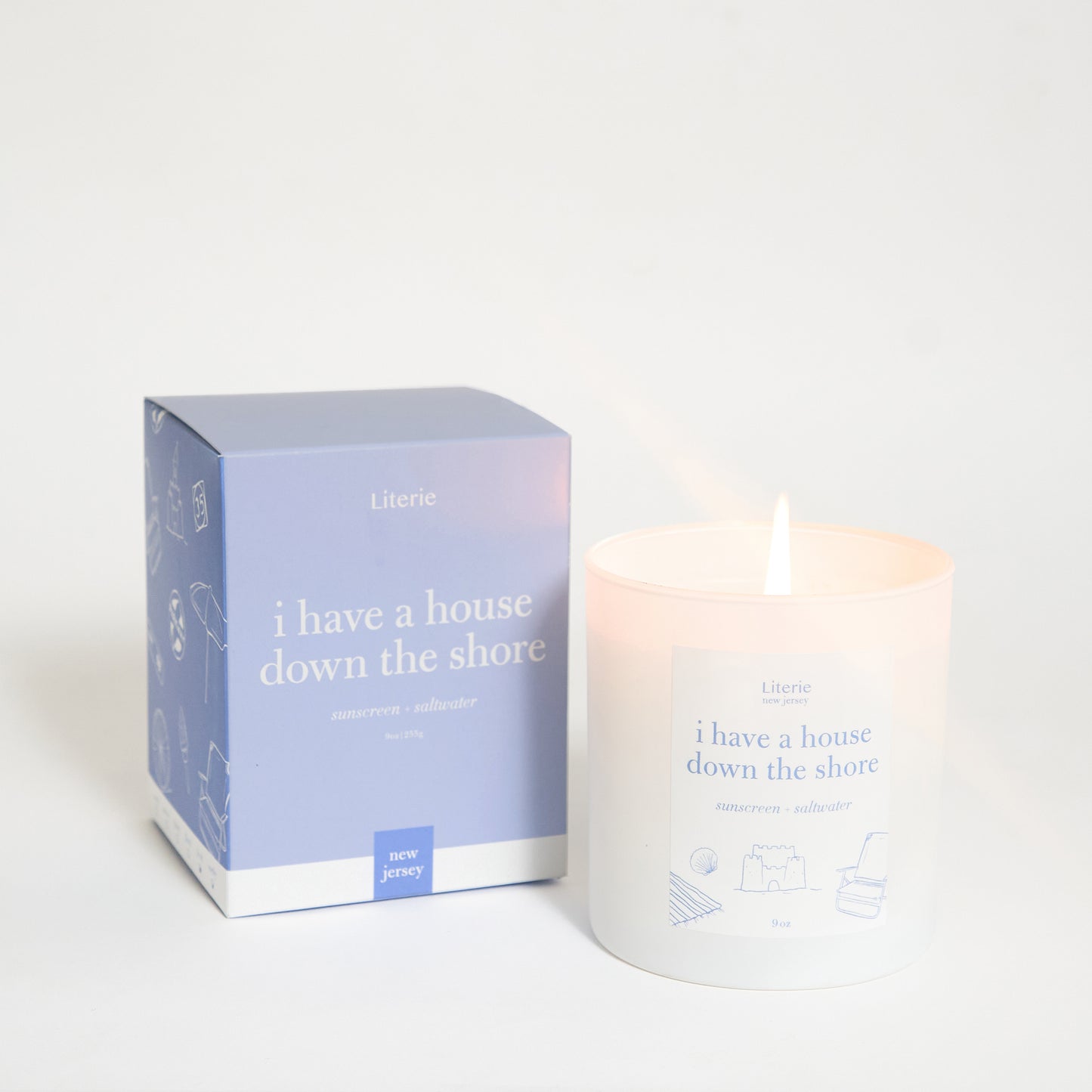 A scented candle with a lit flame, alongside its packaging box which has text and design elements. The candle and box are primarily white and blue.
