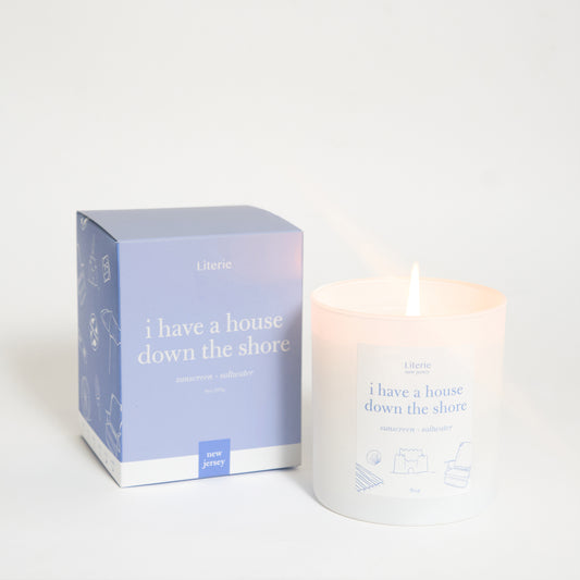A scented candle with a lit flame, alongside its packaging box which has text and design elements. The candle and box are primarily white and blue.