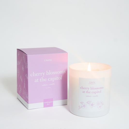 A scented candle with its packaging box, the candle is white and the box is pink with text and floral patterns.
