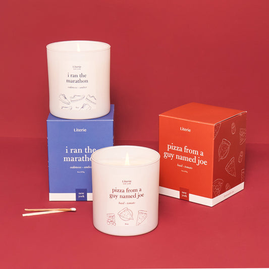 Two candles with labels 'i ran the marathon' and 'pizza from a guy named joe', along with their respective packaging boxes, on a red background.