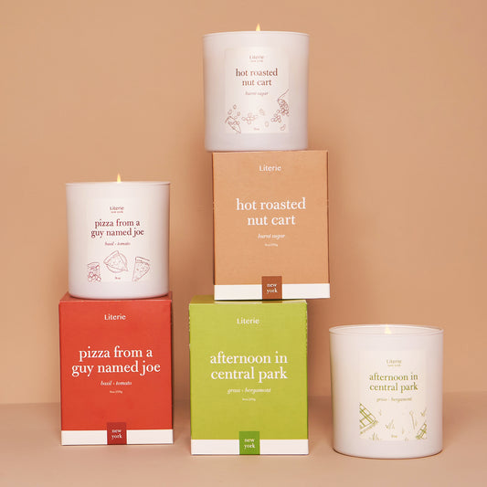 A set of candles with branding and scents related to New York City, presented in white containers with labels in various fonts and colors, stacked on boxes with matching designs.