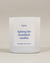 A white glass candle jar with a white rectangular label featuring blue text that reads 'Literie lighting the hanukkah candles' and 'POWDERED JELLY DONUTS' at the bottom, set against a neutral beige background.