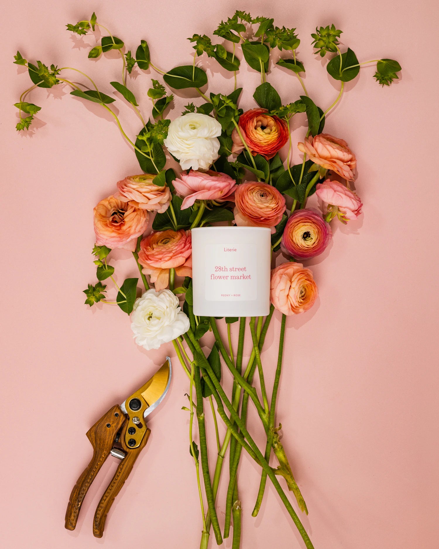 Bouquet of flowers with a '28th street flower market' candle and pruning shears on a pink background.