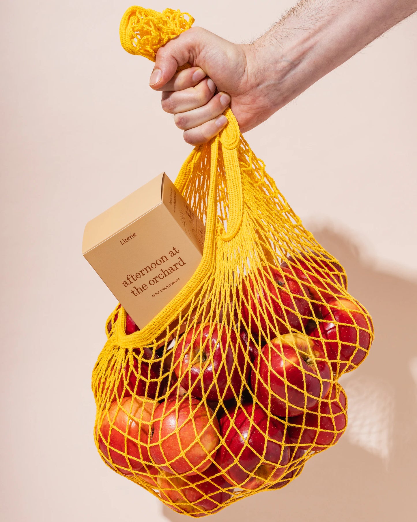 Yellow mesh bag filled with apples and an 'afternoon at the orchard' candle box, held by a hand against a beige background