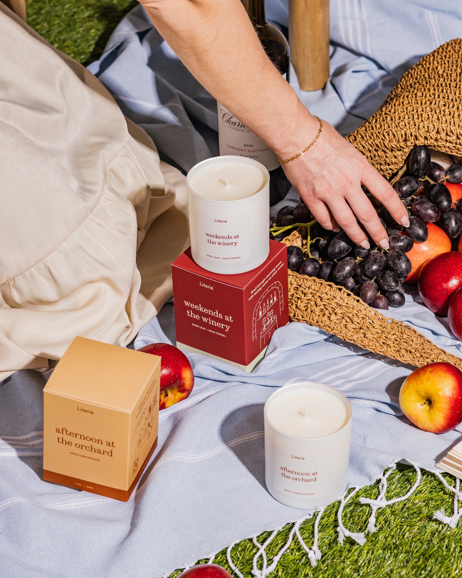 "afternoon at the orchard" and "weekends at the winery" candles and fruit on a picnic blanket with a person's hand reaching for grapes.