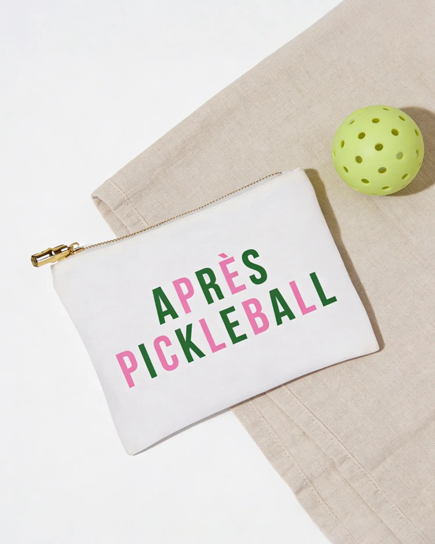 White pouch with 'APRèS PICKLEBALL' text next to a green pickleball ball on a beige surface.