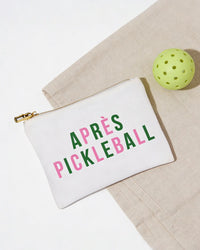 White pouch with 'APRèS PICKLEBALL' text next to a green pickleball ball on a beige surface.