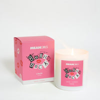 A candle with a pink label and white container, next to its packaging box which has 'Burn Book' text and lipstick print designs.