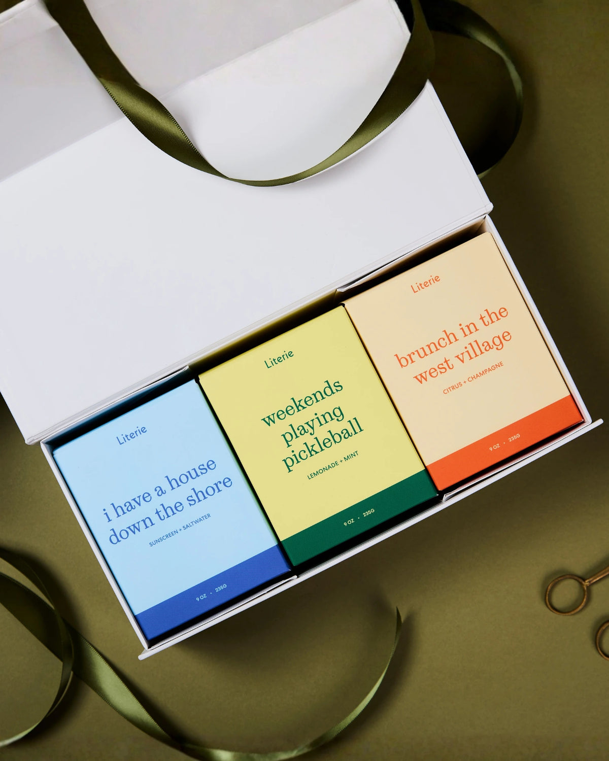 A white gift box containing three colorful candle boxes labeled 'Literie' with playful phrases: 'i have a house down the shore' in blue, 'weekends playing pickleball' in yellow-green, and 'brunch in the west village' in peach-orange. An olive ribbon is draped around the box on a warm brown background.