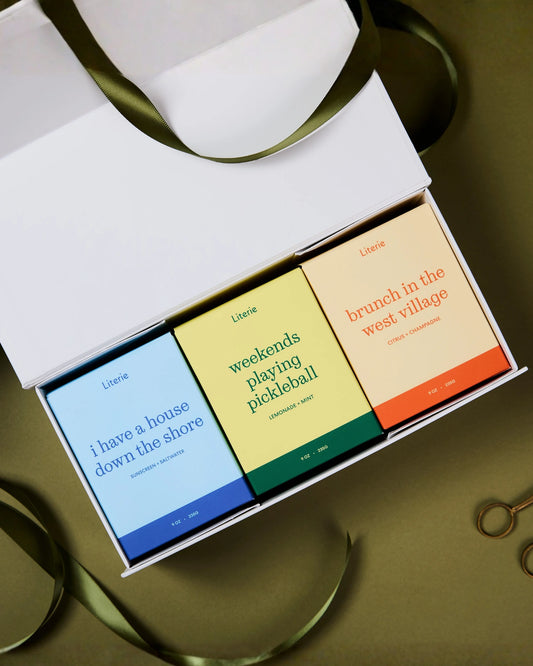 A white gift box containing three colorful candle boxes labeled 'Literie' with playful phrases: 'i have a house down the shore' in blue, 'weekends playing pickleball' in yellow-green, and 'brunch in the west village' in peach-orange. An olive ribbon is draped around the box on a warm brown background.