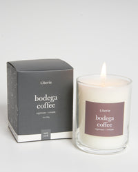 A candle with a lit flame, next to its gray packaging box, labeled 'bodega coffee'.