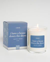 A Literie branded candle with the title 'i have a house down the shore' displayed on its clear glass container, alongside its blue packaging box with the same title.
