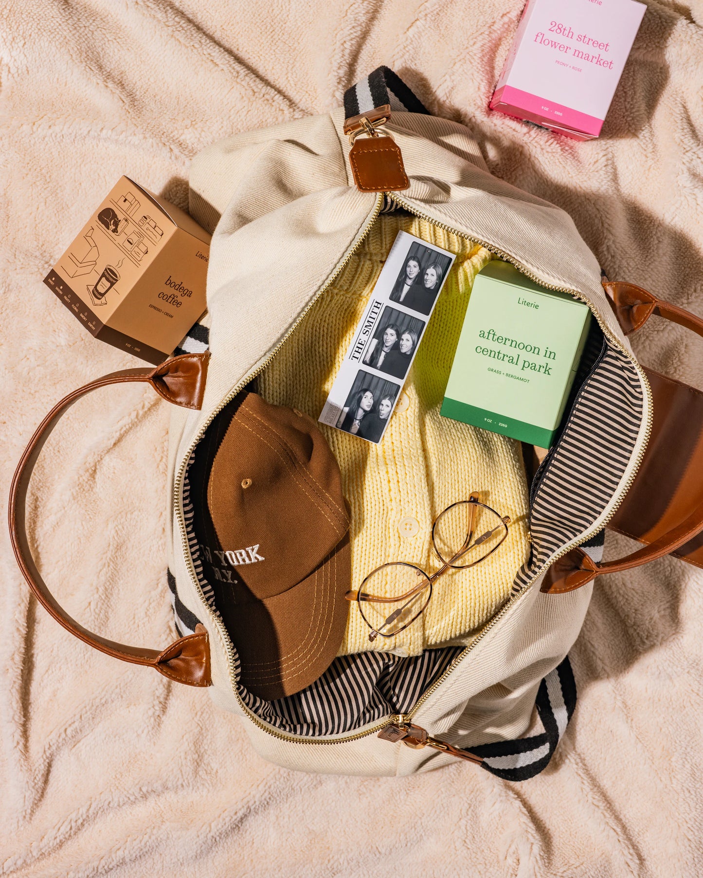 Open beige bag with items including a cap, sunglasses, 28th street flower market, bodega coffee, and afternoon in central park candles on a beige blanket.