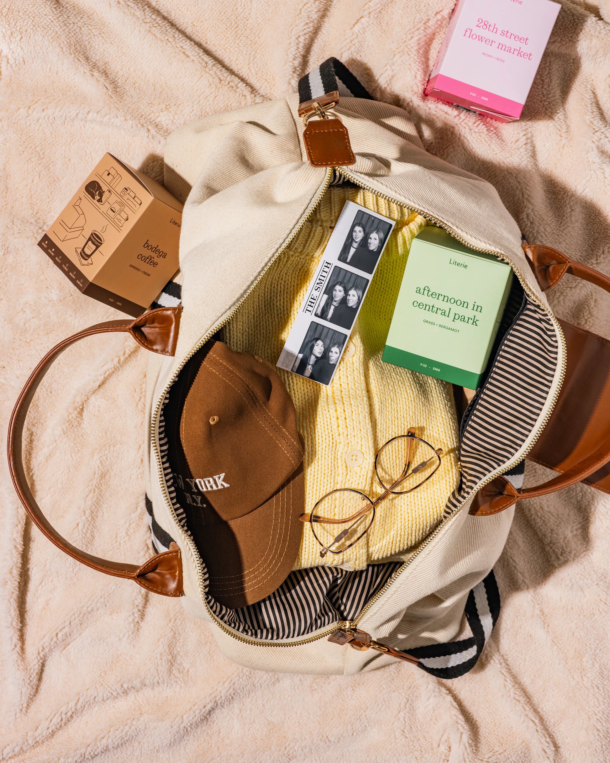 Open beige bag with items including a cap, sunglasses, 28th street flower market, bodega coffee, and afternoon in central park candles on a beige blanket.
