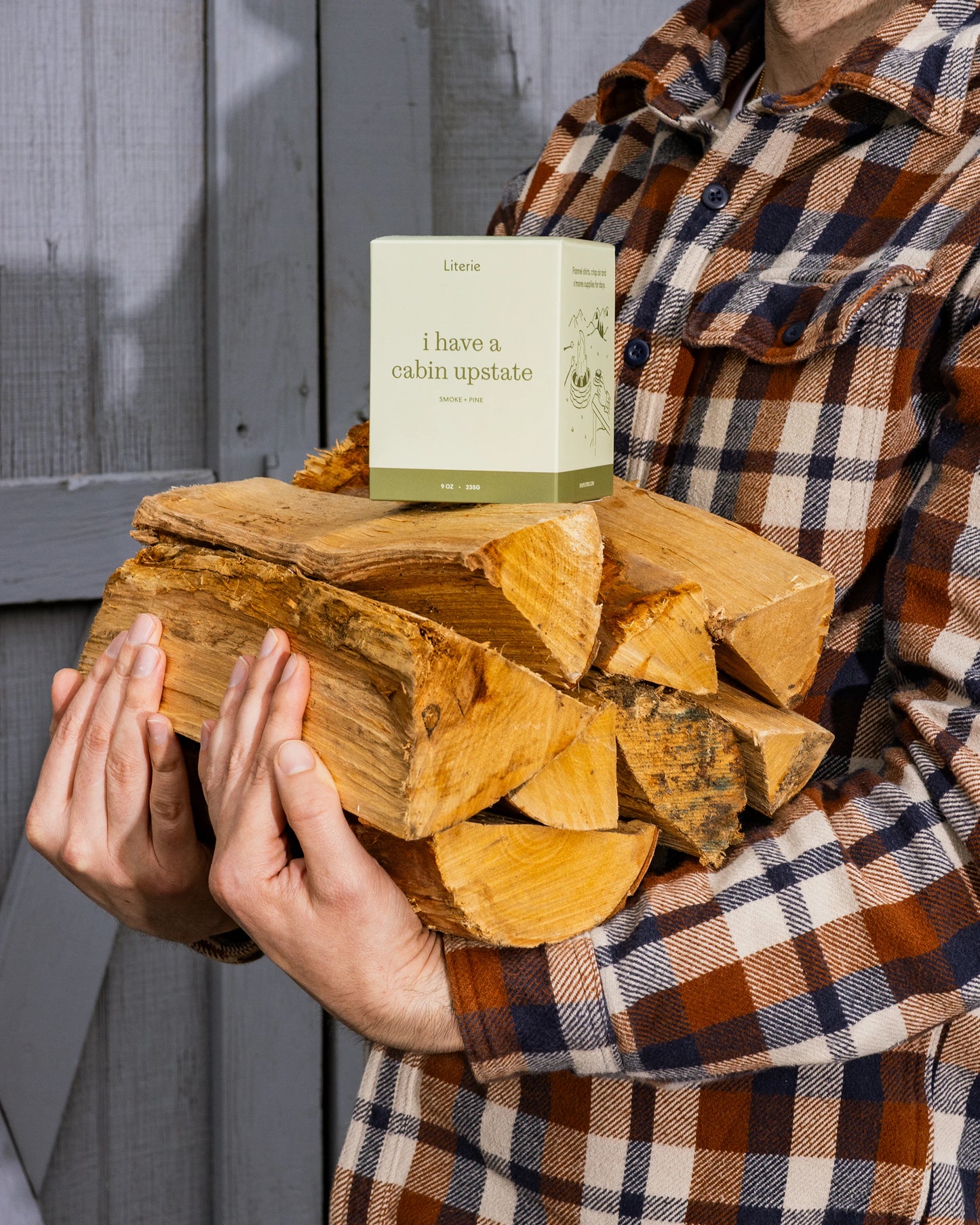 Person holding a stack of firewood with a Literie 'I have a cabin upstate' candle on top.