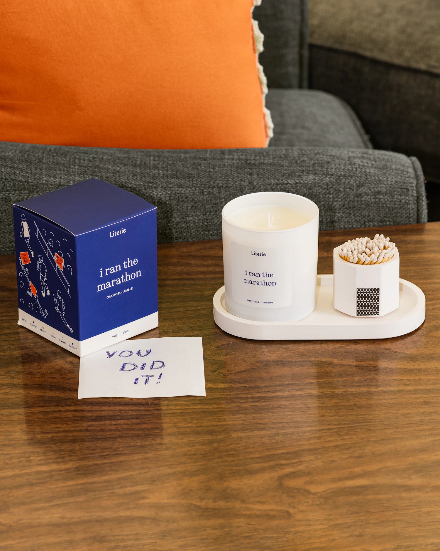 A white candle jar labeled 'I ran the marathon' sits on a white tray with a small match holder and a blue Literie box on a wooden table. An orange pillow and gray sofa are visible in the background.