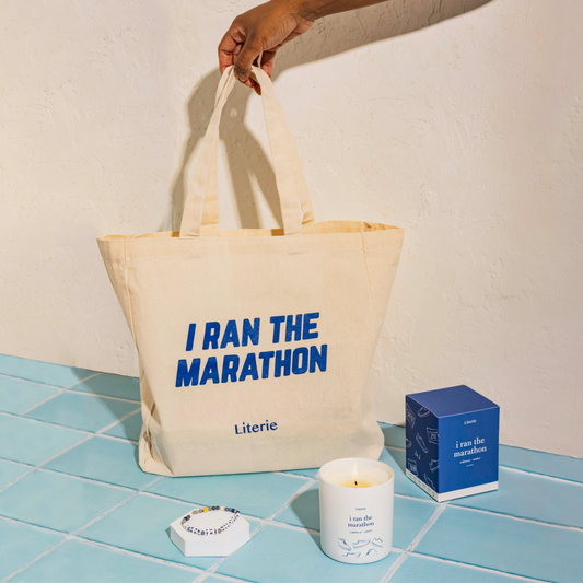 Beige tote bag with 'I Ran the Marathon' text, candle, and box on a tiled floor.