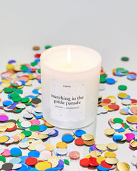 Candle with a label reading 'marching in the pride parade' surrounded by colorful confetti on a light background.