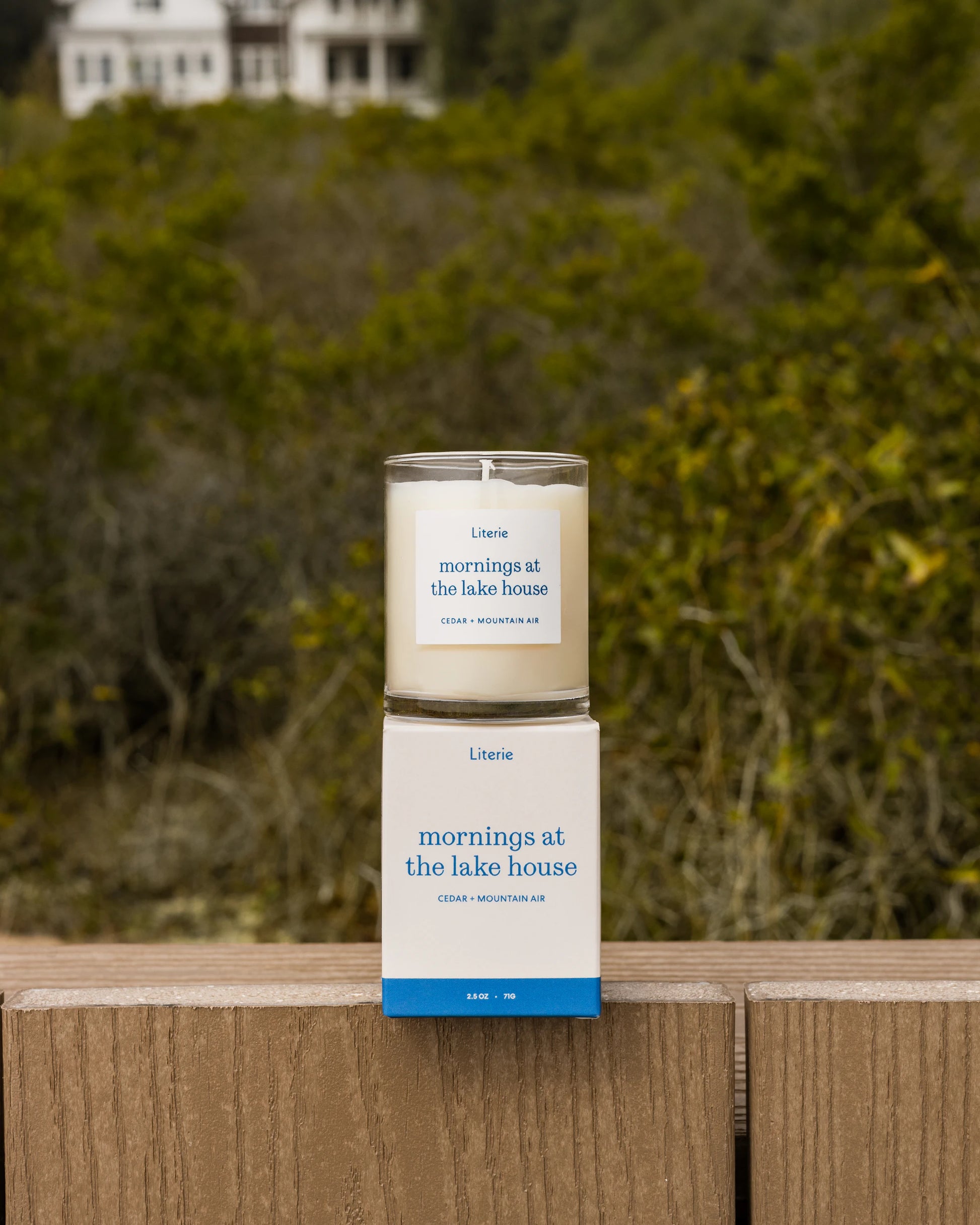 Candle with packaging labeled 'Mornings at the Lake House' on a wooden surface with greenery in the background.