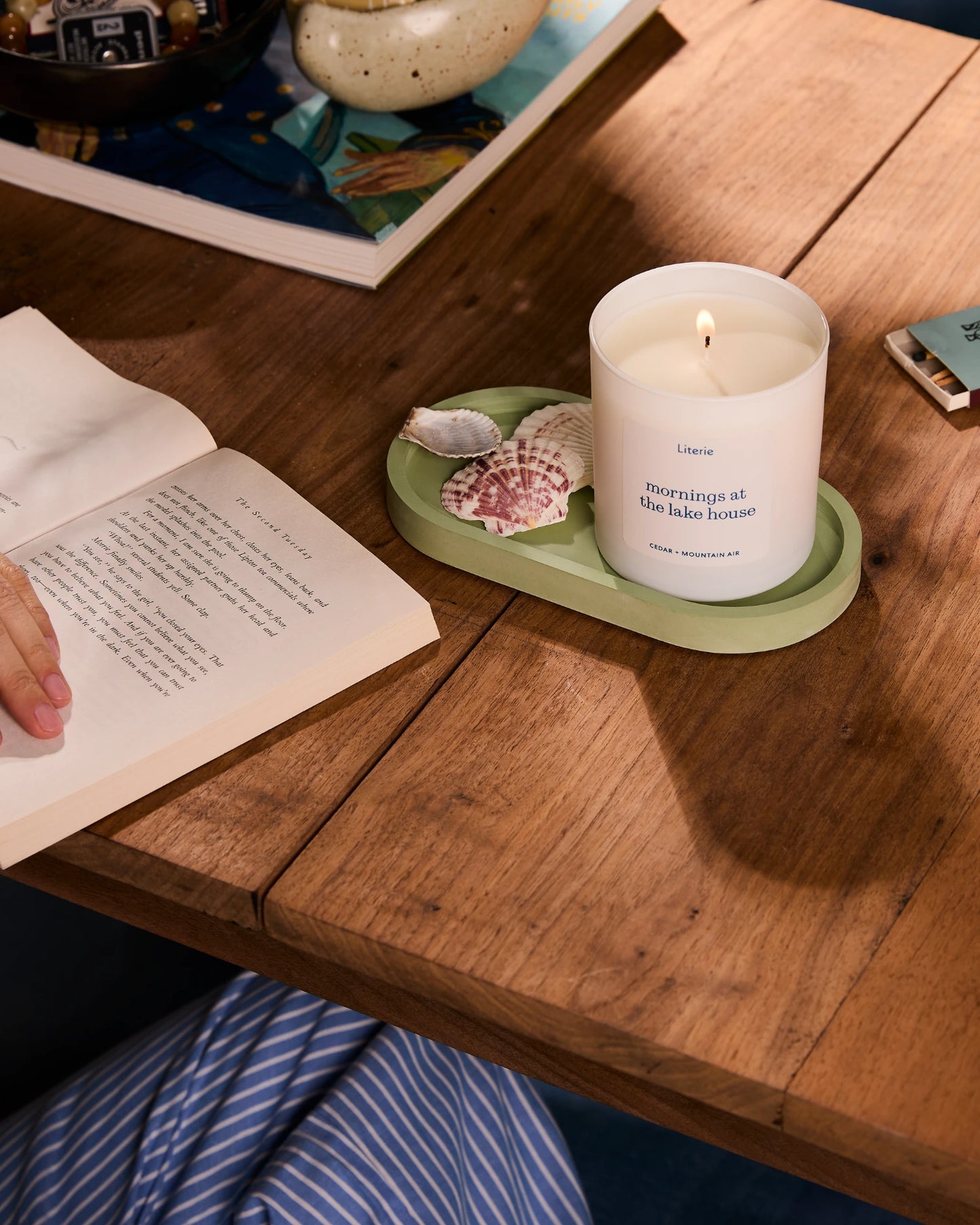 'mornings at the lake house' candle on a wooden table with an open book and seashells nearby.