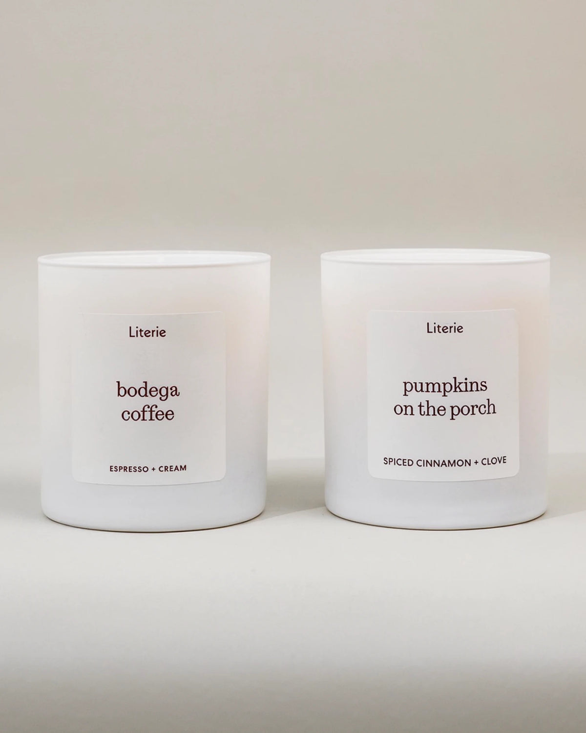 Two candles in white cylindrical jars on a light neutral background. The left label reads 'bodega coffee' with 'ESPRESSO + CREAM' below, and the right label reads 'pumpkins on the porch' with 'SPICED CINNAMON + CLOVE' below.