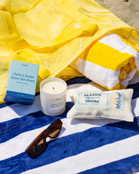 Yellow and blue striped towel with a 'I have a house down the shore' candle, box, and pouch with 'Seaside Shore' text on a beach.
