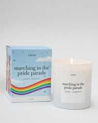 A white candle with text 'marching in the pride parade' on it, alongside a colorful box with rainbow stripes and the name 'Literie'.