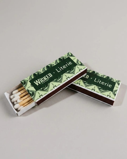 Two WICKED decorative matchboxes with green and white patterned covers and visible wooden matches, photographed on a plain light gray background.