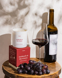 "weekends at the winery" candle and box next to a bottle of red wine, a wine glass, and grapes.
