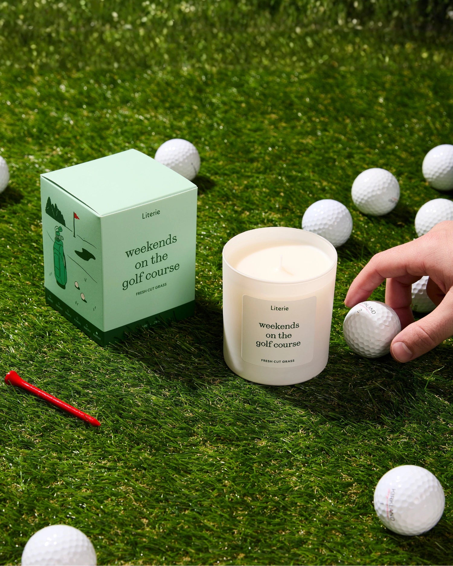 'weekends on the golf course' candle with packaging on grass surrounded by golf balls and a red tee.