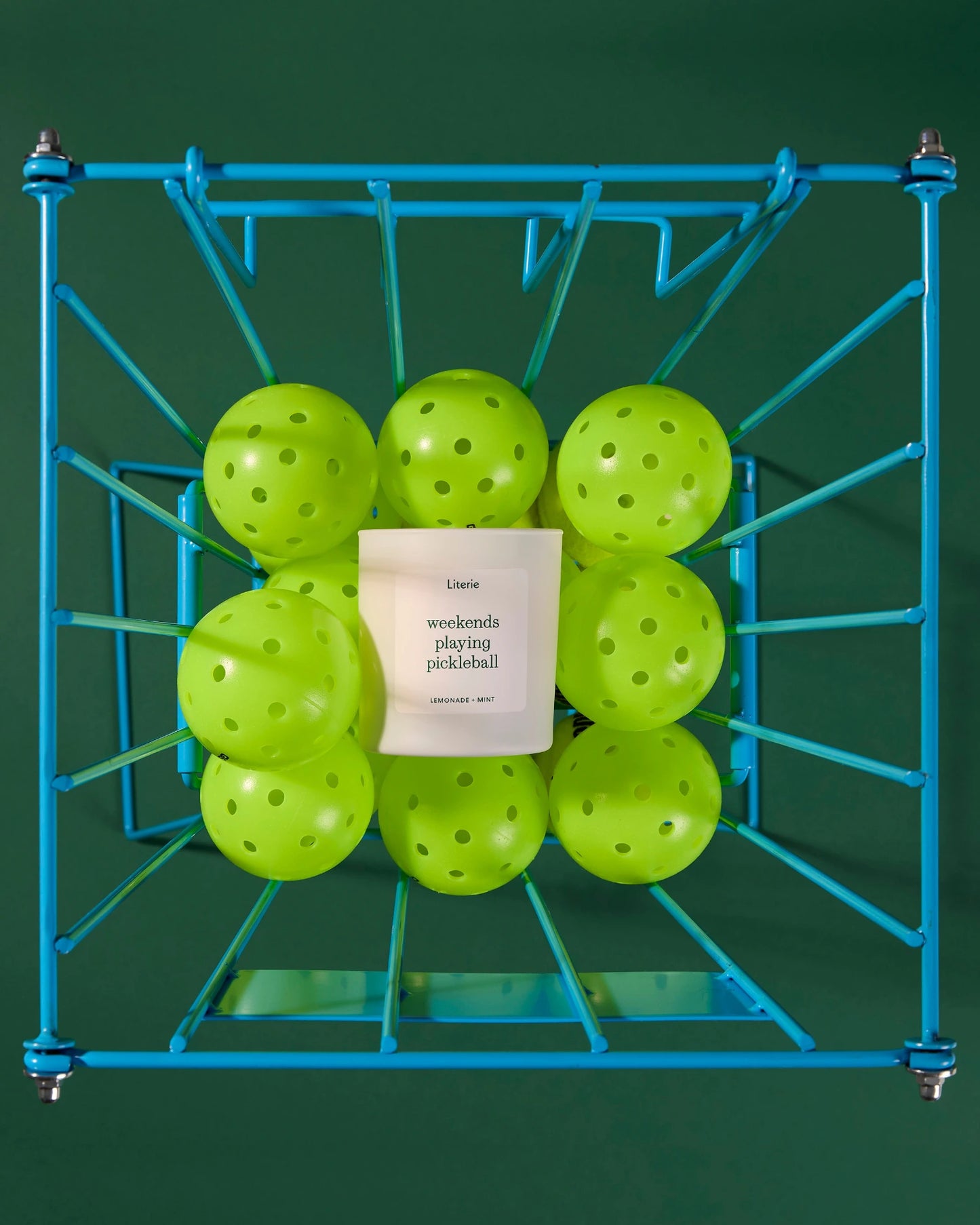 Green pickleballs in a blue metal rack with a 'weekends playing pickleball' candle on a green background