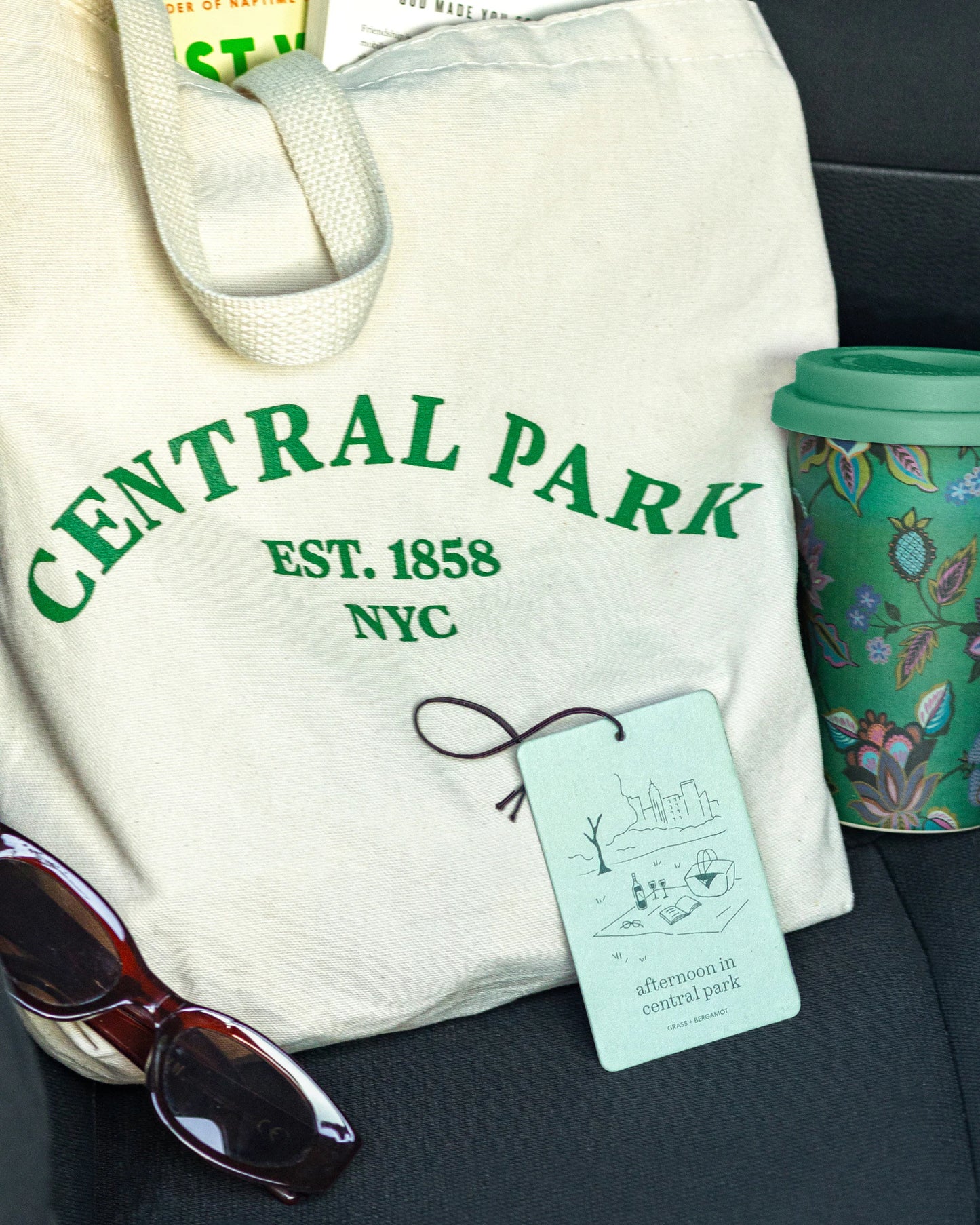 White tote bag with 'Central Park' text, afternoon in centralpark air freshener by Literie, sunglasses, and a mug in a car seat.