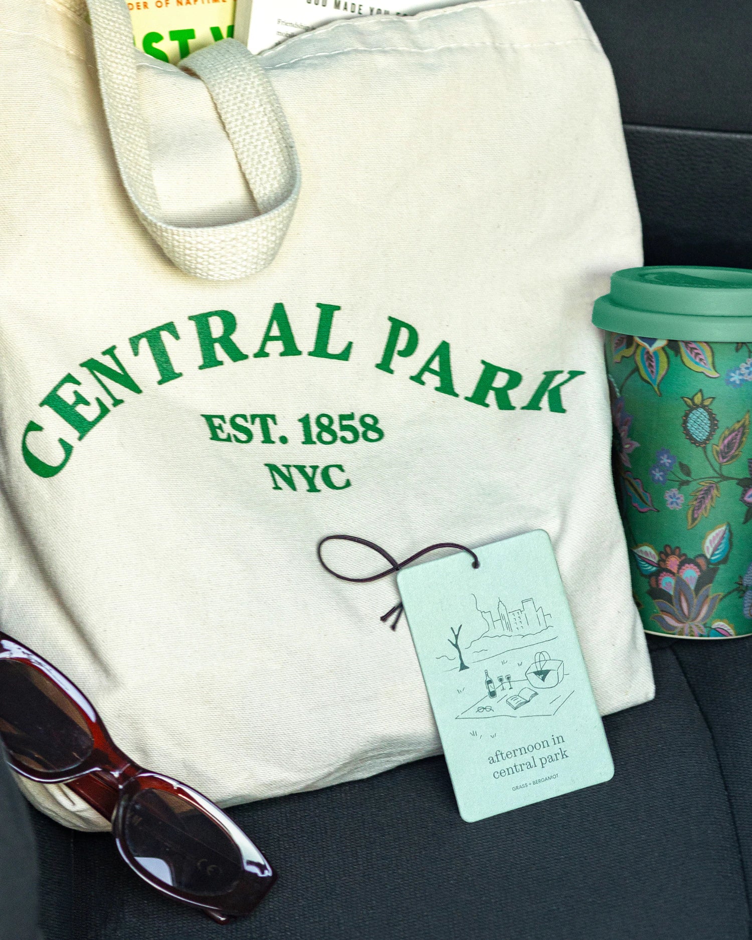 White tote bag with 'Central Park' text, afternoon in centralpark air freshener by Literie, sunglasses, and a mug in a car seat.