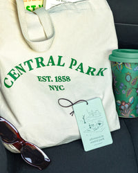 White tote bag with 'Central Park' text, afternoon in centralpark air freshener by Literie, sunglasses, and a mug in a car seat.