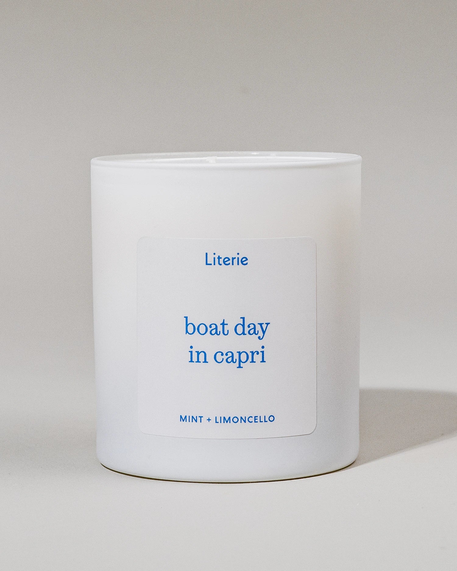 White candle with blue text that says "boat day in capri"and "mint + limoncello" on a light gray background
