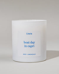White candle with blue text that says "boat day in capri"and "mint + limoncello" on a light gray background