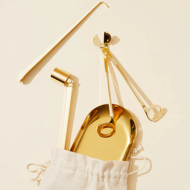 Set of gold candle tools including a tray, scissors, and tweezers on a beige background in a bag.