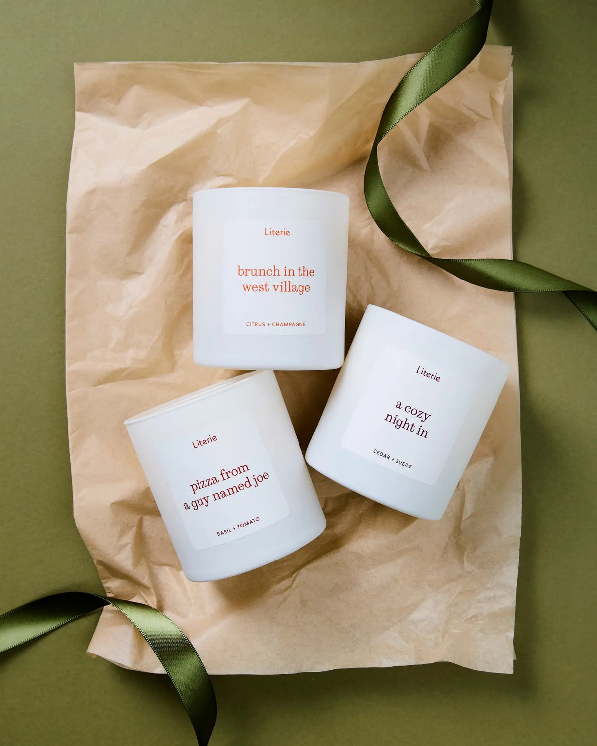 Three white scented candles from the brand Literie are arranged on a sheet of brown kraft paper with a green ribbon, set against an olive green background. The candles feature minimalist labels with red text and are photographed from a top-down angle.
