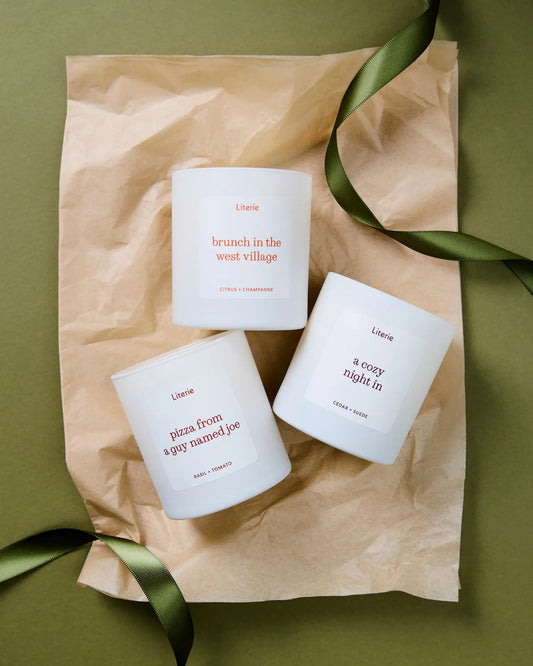 Three white scented candles from the brand Literie are arranged on a sheet of brown kraft paper with a green ribbon, set against an olive green background. The candles feature minimalist labels with red text and are photographed from a top-down angle.