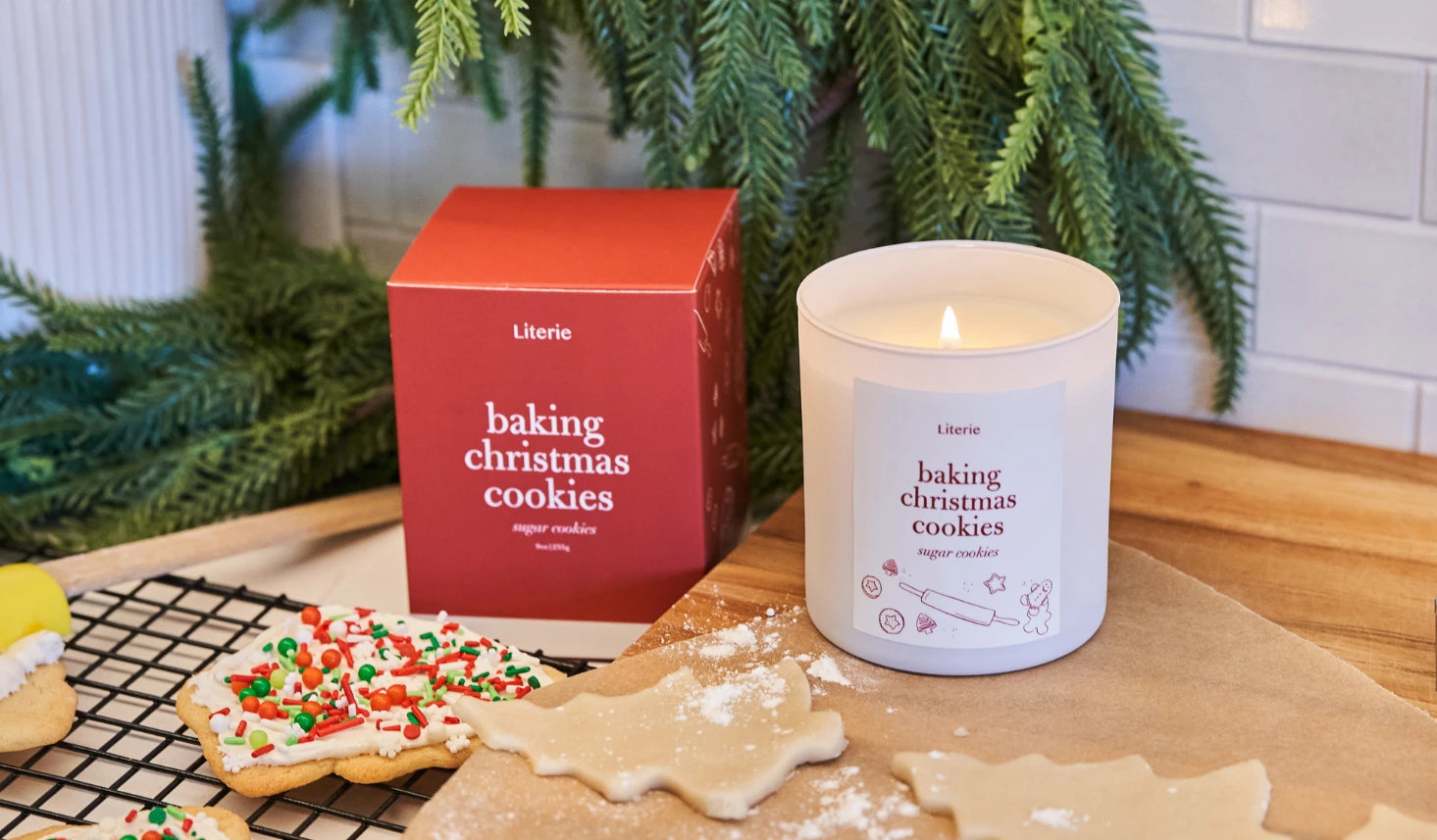 Candle labeled 'baking Christmas cookies' on a table with cookies and a box in a kitchen setting.